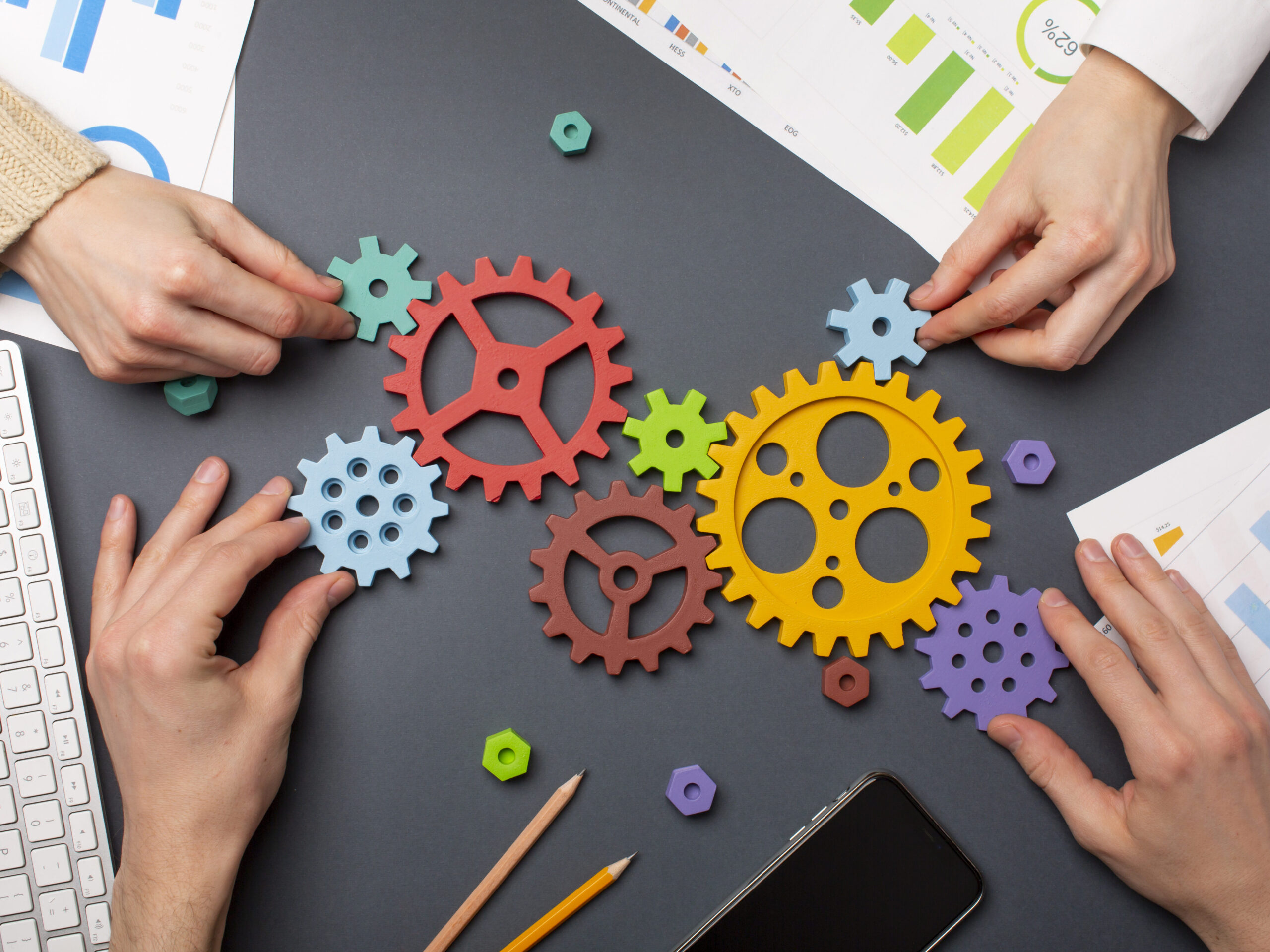 歯車が机の上に並べられ2人の両手が添えられている